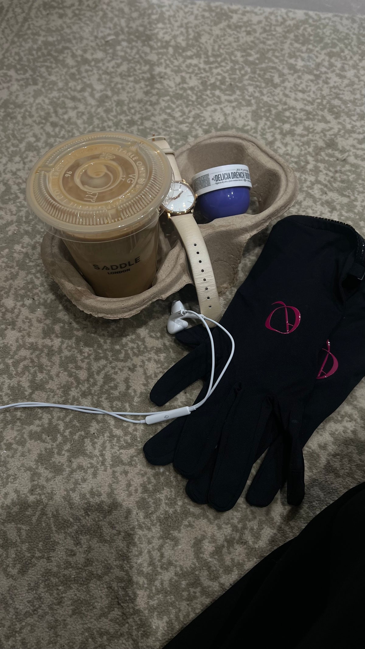 A black knit glove with pink embroidered logo details, displayed alongside a brown paper coffee cup holder, a plastic iced coffee cup, white wired earbuds, and a watch on a textured gray carpet.